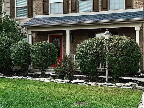 The front of a brick house with four large overgrown shrubs blocking the porch, and a white lamp post standing in a flagstone-edged bed.
