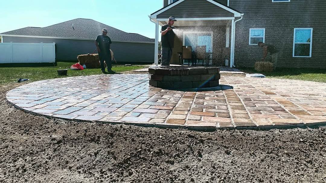 A newly finished circular paver patio with a stone fire pit at its centre, laid behind a two-storey house, with two of the crew still at work.