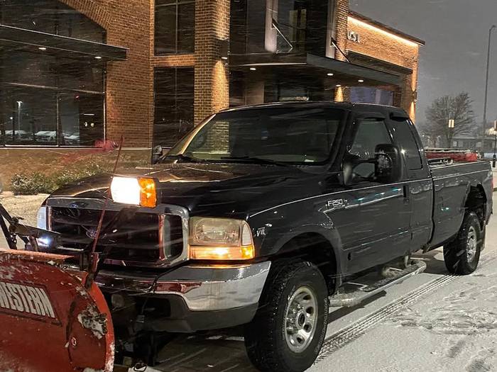 A black pickup with a red snow plow mounted on the front, parked on a cleared commercial lot at night with snow on the ground.